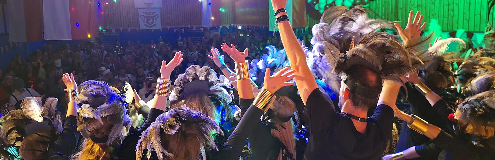 Blick von der Bühne auf eine Tanzgruppe mit Federschmuck und verkleidetes Publikum in einer Festhalle während einer Karnevalsveranstaltung